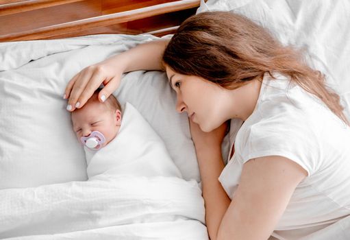 Beautiful young mother lying in the bed with newborn daughter and looking at her holding hand on child head. Adorable infant baby girl sleeping close to her mom parent