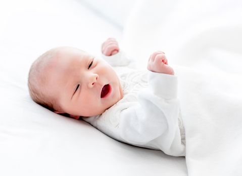 Closeup portrait of adorable newborn baby girl wearing white costume lying in the bed with mouth open. Cute infant child resting at home