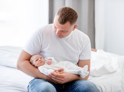 Adorable newborn baby girl wearing white costume sleeping in father hands in bedroom with daylight. Cute infant child napping at home and parent dad holding her