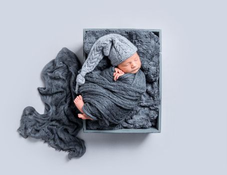 Cute newborn covered with huge dark blanket resting in a basket, wearing big hat, topview