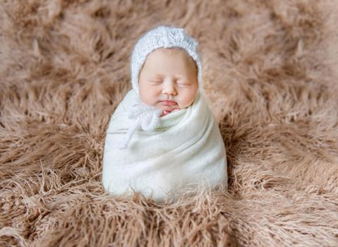 Adorable child in a white hat, wrapped with white blanket, sleeping tightly on furry surface