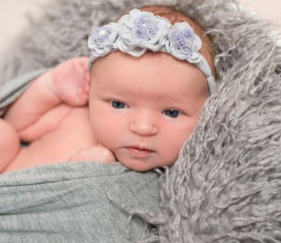 Adorable baby in gray wrap trying to crawl out of it, flowery hairband, furry pillow