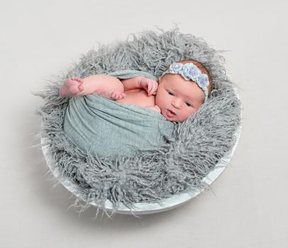 Adorable baby in gray wrap trying to crawl out of it, flowery hairband, furry pillow