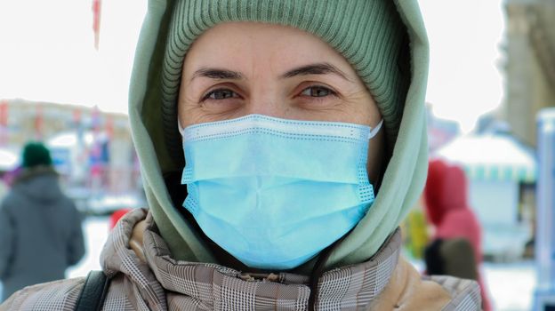Portrait of happy young woman wearing medical protective mask outdoors and looking at camera. Virus protection. A woman outdoors on a winter day stands in a hat and a hood in the park