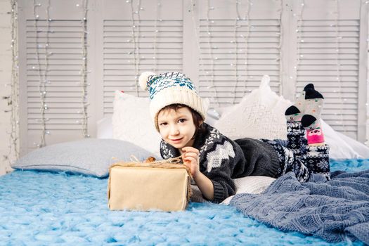 little girl the child sitting in pajamas and hat on the bed with garland of light bulbs with gifts boxes wrapped in a non-colored paper decorated with cones on blue knitted coverlet.Christmas concept.