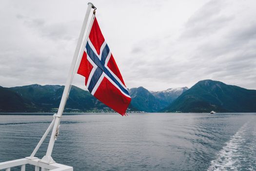 Norwegian flag hanging on the railing of the ship and waving above the water. Norvegian fjord with a flag. Ferry trip in Norway. Norway Flag on sea and mountains background.