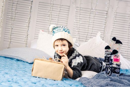 little girl the child sitting in pajamas and hat on the bed with garland of light bulbs with gifts boxes wrapped in a non-colored paper decorated with cones on blue knitted coverlet.Christmas concept.