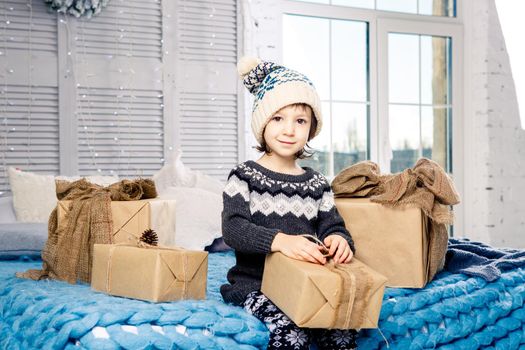 little girl the child sitting in pajamas and hat on the bed with garland of light bulbs with gifts boxes wrapped in a non-colored paper decorated with cones on blue knitted coverlet.Christmas concept.