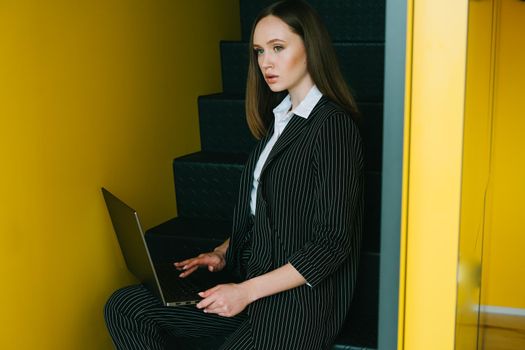 A beautiful girl sits on the stairs and works behind a laptop.