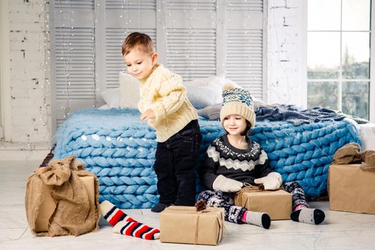 Children brother and sister sit on the floor in bedroom near the bed with gifts on the background of Christmas decor on a sunny day. Dressed in a warm knitted woolen garment cap and colored mittens.