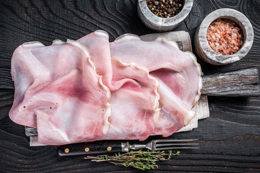 Sliced prosciutto ham on wooden board with herbs. Black wooden background. Top view.