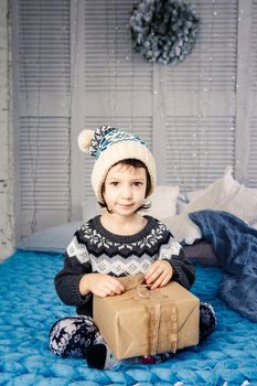 little girl the child sitting in pajamas and hat on the bed with garland of light bulbs with gifts boxes wrapped in a non-colored paper decorated with cones on blue knitted coverlet.Christmas concept.