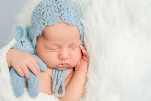 sleepy newborn boy in blue knitted hat with toy on white fluffy blanket