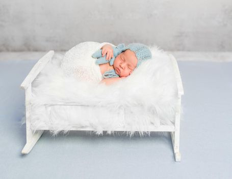 dreamy newborn boy holding toy sleeps on little bed covered with white fluffy blanket