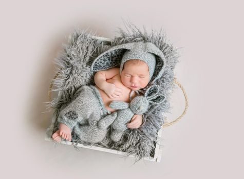 Funny newborn boy in grey bunny bonnet and grey knitted pants sleeping and holding bunny toy in his hand on wooden box with fluffy banket. Top view.