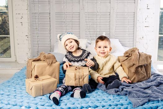 Theme Christmas morning. Two child Caucasian boy and girl brother and sister are sitting on the bed in an embrace with a smile and joy are opening New Year's gifts in boxes on a sunny day.