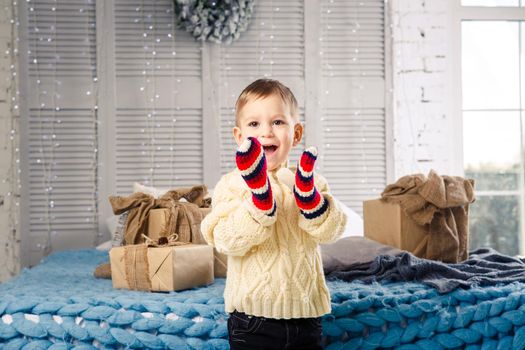 little funny playful boy a child sits on a bed on Christmas day with gift boxes in white wool knitted sweater and big bright mittens on it and laughs out loud. In interior there is a festive decor.