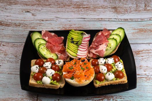 Assorted sandwiches with fish, cheese, meat and vegetables on a black plate and wooden background.