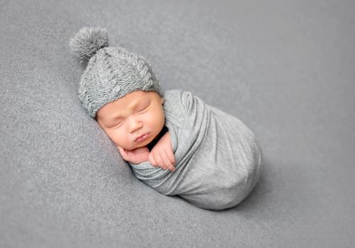 Beautiful newborn baby sleeping curled up in grey blanket and wearing grey hat. Little baby lying on grey surface