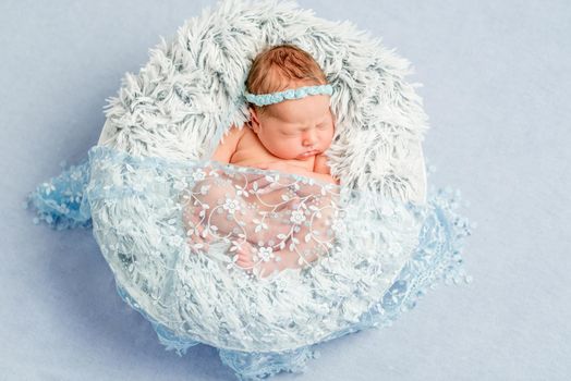 funny newborn girl with headband with flowers in round basket on furry blanket covered with veil