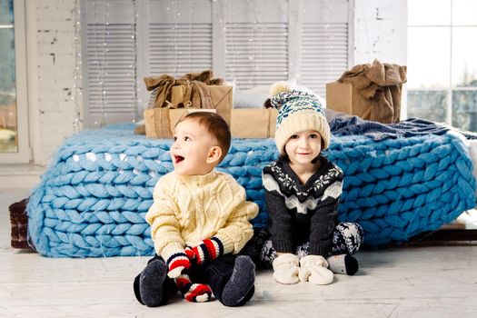 Children brother and sister sit on the floor in bedroom near the bed with gifts on the background of Christmas decor on a sunny day. Dressed in a warm knitted woolen garment cap and colored mittens.