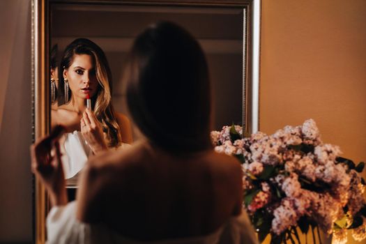A beautiful girl paints her lips in the reflection of the mirror at home. A girl looks in the mirror before the holiday and holds a lipstick in her hands. Girl with long hair goes to a party.