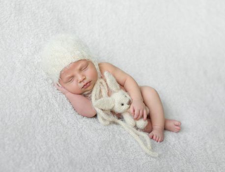 bare sleeping baby in hat with toy on white soft blanket with hand under his head