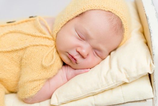 Cute baby dressed in knitted yellow costume sleeping on small wooden crib with one leg on the floor