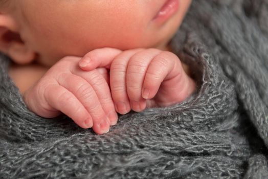 Adorable fingers of a newborn, touching his cheek in his sleep, wrapped in blanket, closeup