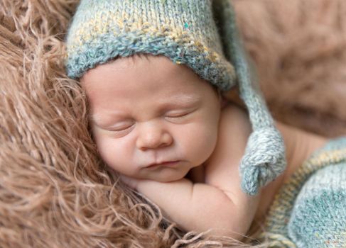Little baby weared in light blue-yellow knitted beanie and pants sweetly sleeping on the light brown soft coverlet