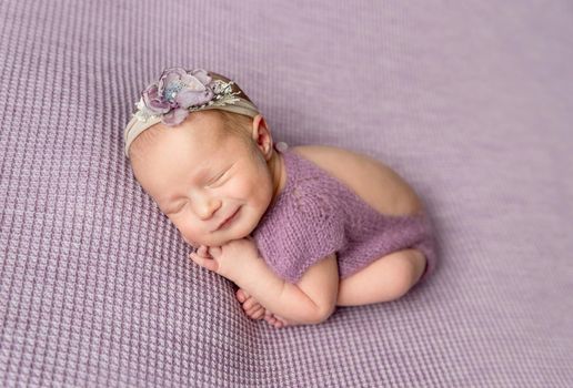 Newborn baby girl smiles in sleep. Little girl in purple bodysuit and headband curled up on blanket
