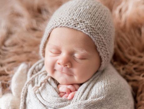 Little cute baby weared in gray knitted beanie and covered with gray knitted blanket sweetly sleeping with bunny nearby on the soft light brown bedcover