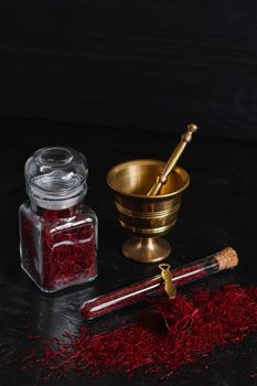 Raw organic red dried saffron spice on wooden background in vintage metal brass mortar with pestle, glass jar and tube.