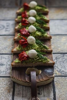 Mini triangle canapes bruschetta with avocado, mozarella and dried tomatos on the rustic board on the vintage tiled background.
