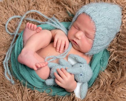 Infant in a cute blue hat napping with his elephant toy on a furry pillow, closeup
