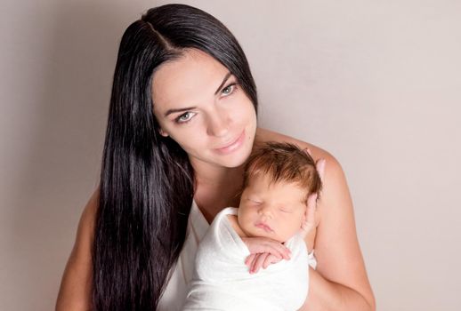 Mother holding little sleeping baby covered in white coverlet on the light background