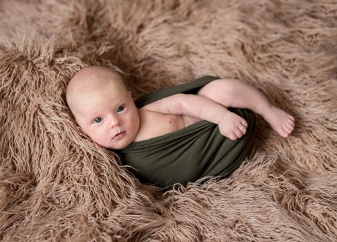 Little cute blue-eyes baby covered in a dark green blanket lying on the soft light brown bedcover