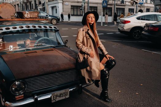 A stylish young woman in a beige coat and black hat on a city street sits on the hood of a car at sunset. Women's street fashion. Autumn clothing.Urban style.