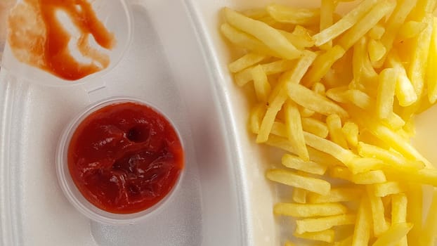 A close-up of golden orange yellow deep fries with tomato ketchup served in a foam container. Fast food takeaway. Unwanted fresh food
