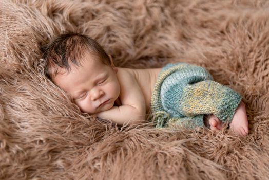 Little baby weared in light blue-yellow knitted beanie and pants sweetly sleeping on the light brown soft coverlet