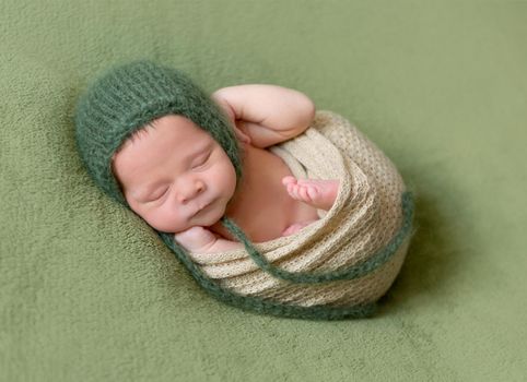 Little baby in dark green knitted beanie covered in lilac blanket sweetly sleeping on the soft green bedcover