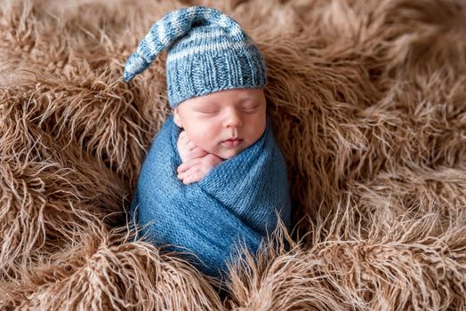 Little cute baby in a light blue knitted beanie and covered with blue knitted blanket sweetly sleeping on the soft light brown bedcover