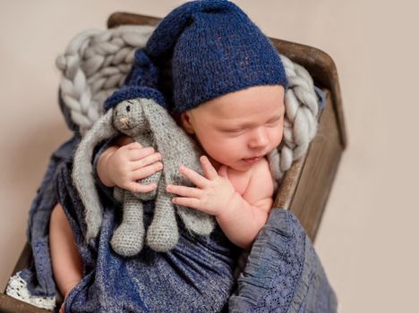 Little smiling baby weared in a blue knitted beanie sweetly sleeping in the small brown bed covered with blue-gray blankets