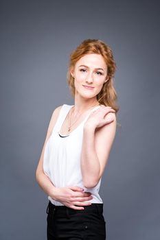 portrait of a young red-haired beautiful girl in the studio on a gray isolated background. A woman is standing with her arms folded and smiling in a white shirt with a short sleeve. Business concept