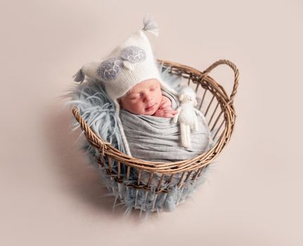 Side view of baby in white knitted hat with toy sleeping in basket covered in blanket