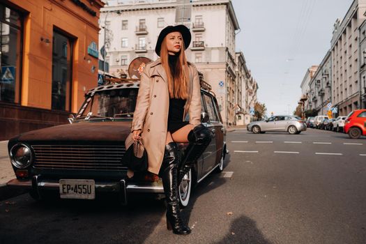 A stylish young woman in a beige coat and black hat on a city street sits on the hood of a car at sunset. Women's street fashion. Autumn clothing.Urban style.