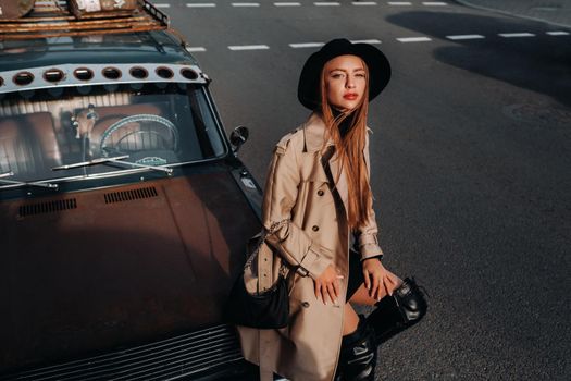 A stylish young woman in a beige coat and black hat on a city street sits on the hood of a car at sunset. Women's street fashion. Autumn clothing.Urban style.