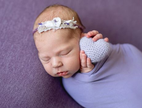 Sleepy beatiful newborn child with knitted heart-shaped toy on violet blanket