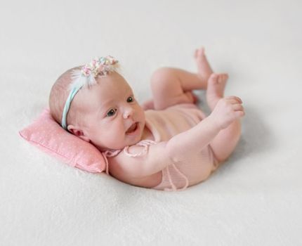 Little smiling baby in pink knitted suit joyfully lying on pink dotted pillow and white soft blanket