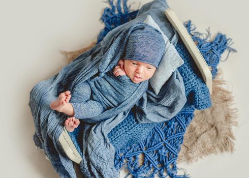 Sweet newborn baby in blue clothing sleeps in a little bed on knitted carpet